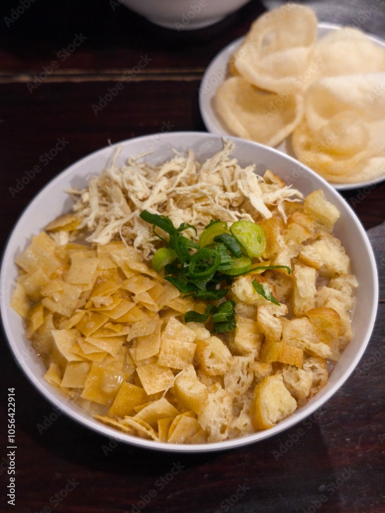 A bowl of warm chicken porridge paired with delightful blurry shrimp crackers. Indonesian food consist of white rice porridge, shredded chicken, cakwe, crackers, and leek. A bowl of warm chicken porridge paired with delightful blurry shrimp crackers. Indonesian food consist of white rice porridge, shredded chicken, cakwe, crackers, and leek.