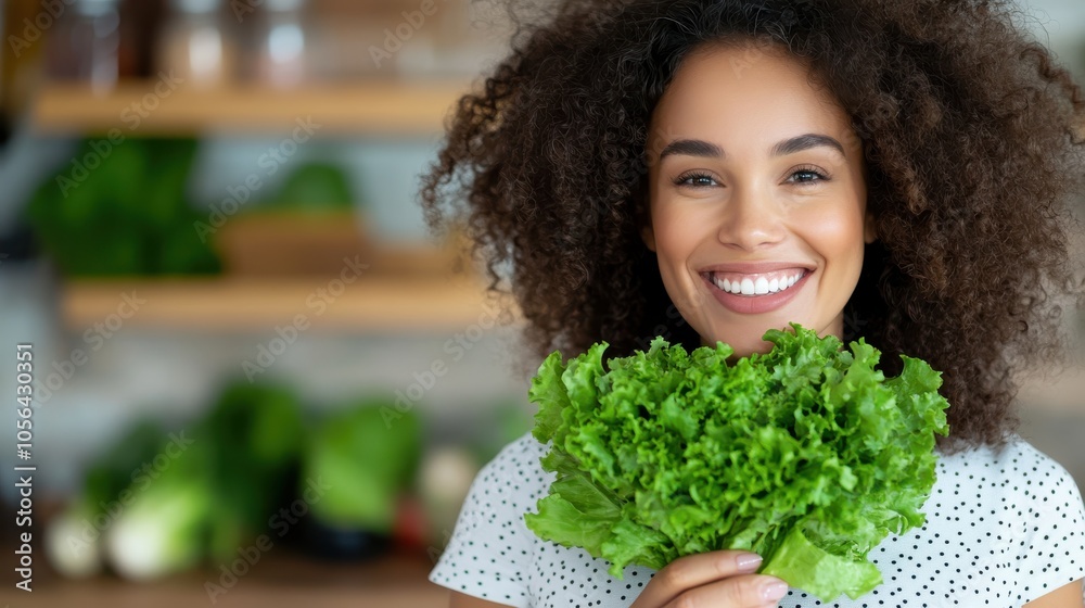 A cheerful woman with curly hair holds fresh green lettuce, symbolizing health and wellness in her modern kitchen with various vegetables in the background. A cheerful woman with curly hair holds fresh green lettuce, symbolizing health and wellness in her modern kitchen with various vegetables in the background.