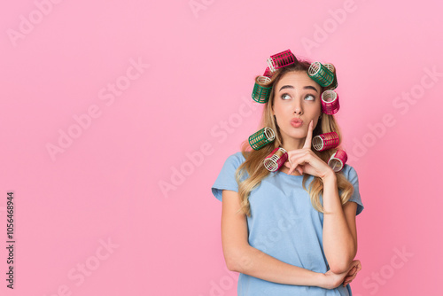 Pensive young housewife in curlers presses finger to cheek and looks up to empty space, isolated on pink background, studio shot