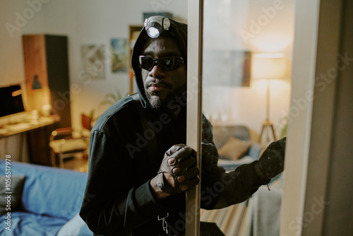 African American thief with headlamp opening cupboard carefully while glancing around