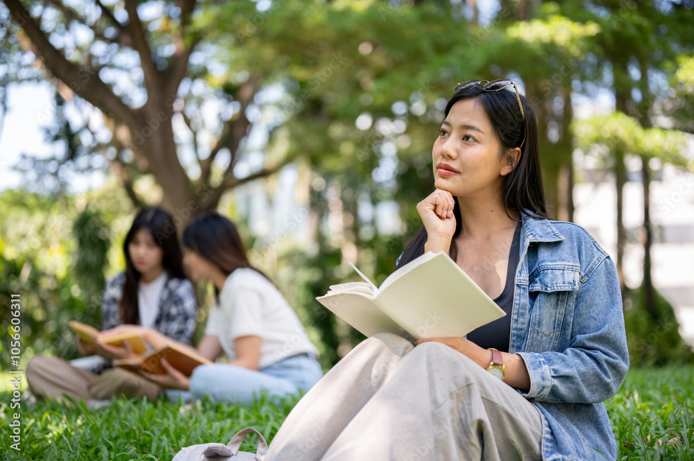 Obraz premium An attractive Asian woman sits on grass in a college campus park, daydreaming while reading a book.