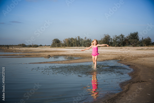 A little seven-year-old girl in a pink swimsuit runs along the lake in a good cheerful mood with a smile on her face. Summer lifestyle. Health tempering with water procedures and outdoor walks. photo