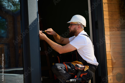 Construction worker installing door in a new house construction site. Man in working attire install front door. Builder in uniform with screwdriver adjusting front glass door in new beautiful house.