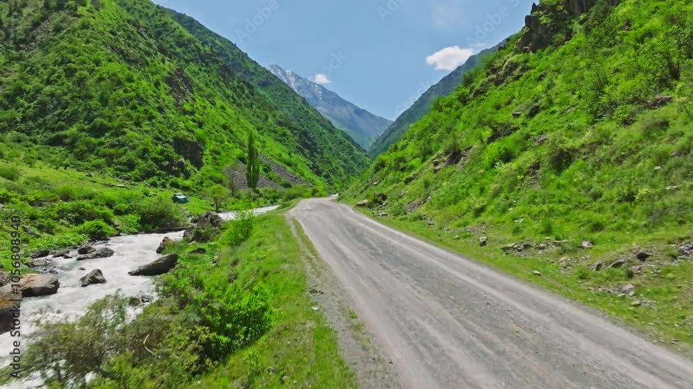 flying along mountain river in narrow mountain valley with empty dry dirt road and green slopes at sunny summer day in Kyrgyzstan