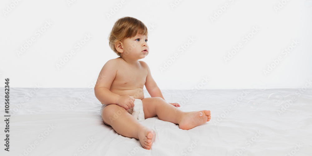 Side profile of curious baby, toddler sitting in diaper on soft bed, gazing upwards with joy against white studio background. Concept of parenthood, natural beauty, love and care. Ad. Side profile of curious baby, toddler sitting in diaper on soft bed, gazing upwards with joy against white studio background. Concept of parenthood, natural beauty, love and care. Ad.