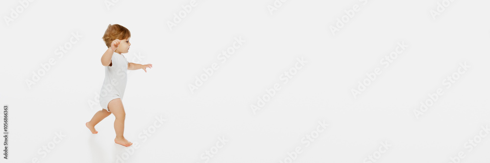 Banner. Lively, playful toddler running in onnesie, joyfully raising hands against white studio background with negative space for text. Concept of parenthood, natural beauty, love and care. Ad. Banner. Lively, playful toddler running in onnesie, joyfully raising hands against white studio background with negative space for text. Concept of parenthood, natural beauty, love and care. Ad.