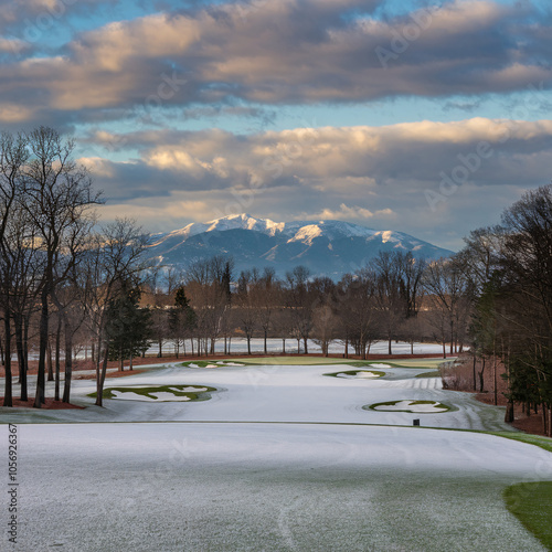 A winter golf course features snow-covered fairways, bare trees, and snow-capped mountains under a cloudy sky.