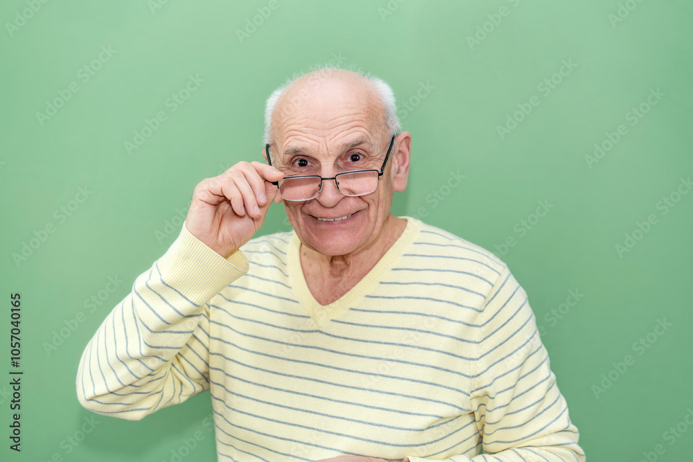 Old man smiling warmly, lifting his glasses while looking at camera against green background, wearing striped yellow sweater Old man smiling warmly, lifting his glasses while looking at camera against green background, wearing striped yellow sweater