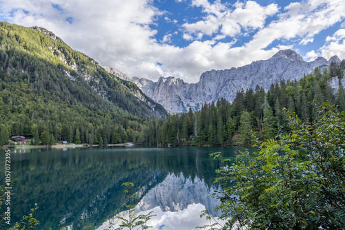 vista panoramica del lago di Fusine inferiore visto da una delle sponde, con la catena montuosa del monte Mangart a distanza, che si riflette sull'acqua insieme alla vasta foresta photo