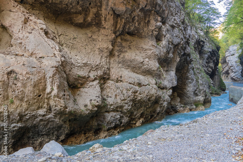 Steep cliffs hang over mountain river with emerald-clear water. Stormy Chegem River flows in deep crevice along bottom of Chegem Gorge. In depths of gorge, semi-darkness reigns.