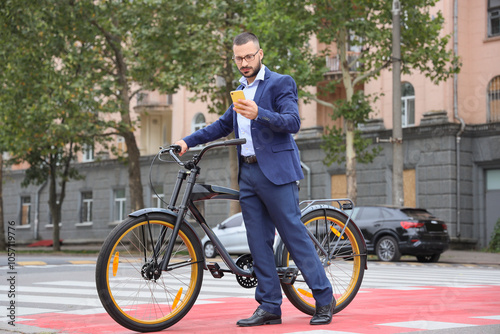 Young businessman with mobile phone and bicycle crossing road on city street