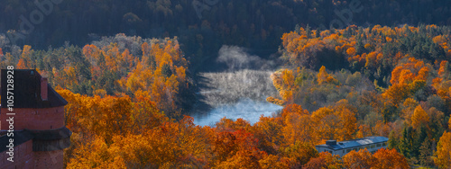 The image shows Turaida Castle's red brick tower surrounded by golden autumn trees in Sigulda. A misty river winds through the vibrant landscape. photo