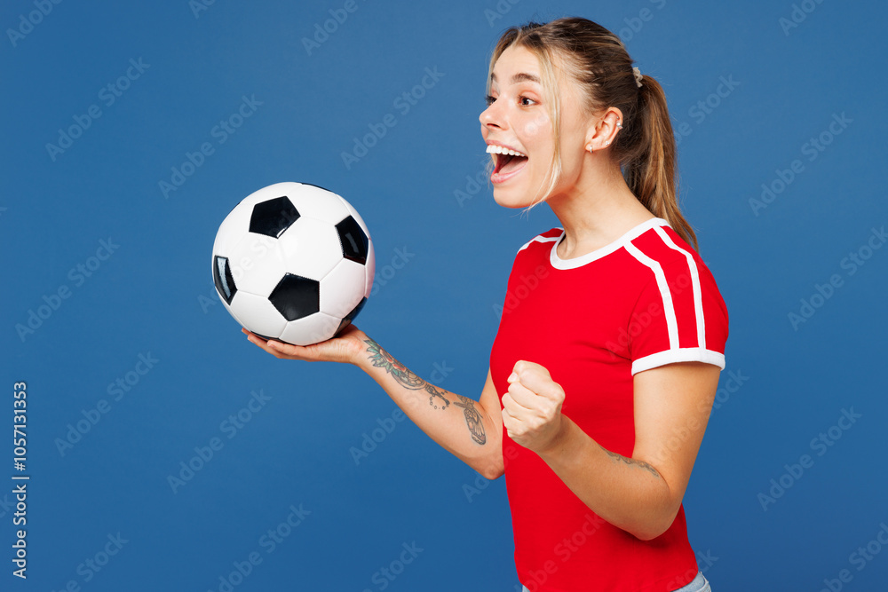 Φωτογραφία Young woman fan wears basic red t-shirt cheer up support football sport team hold in hand soccer ball watch tv live stream doing winner gesture celebrate clench fist isolated on plain blue background