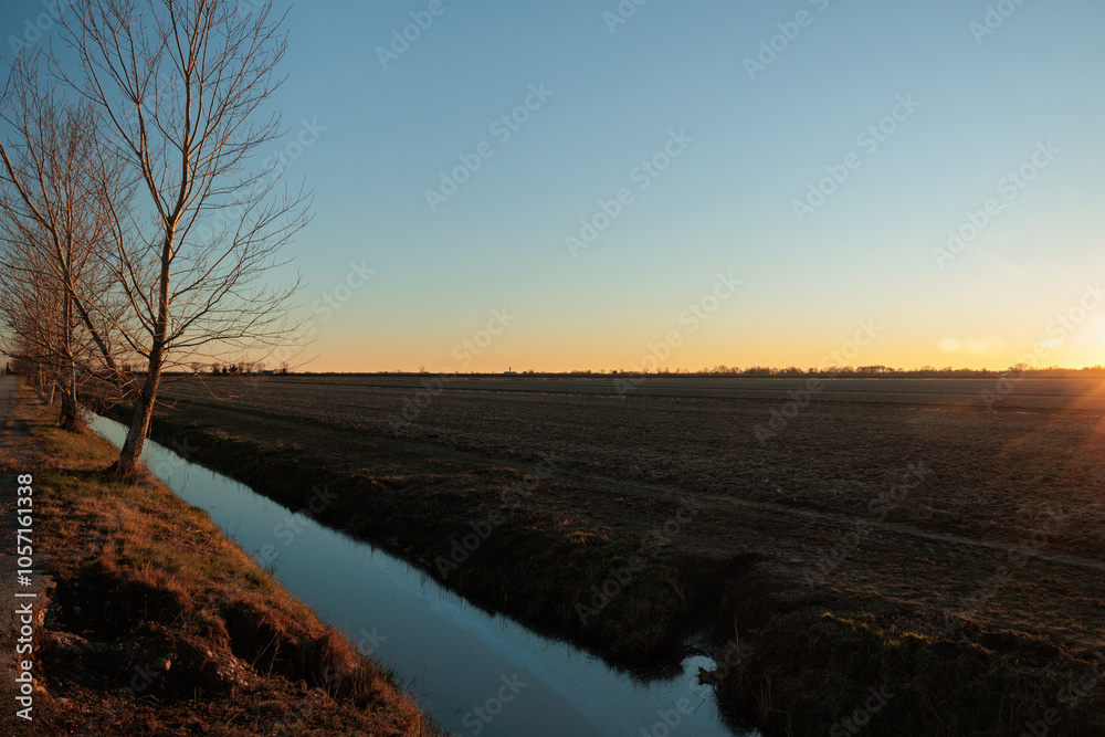 sole che tramonta sulla linea dell'orizzonte e colora di arancione il cielo azzurro e sereno, con vista panoramica da un ambiente di campagna in autunno