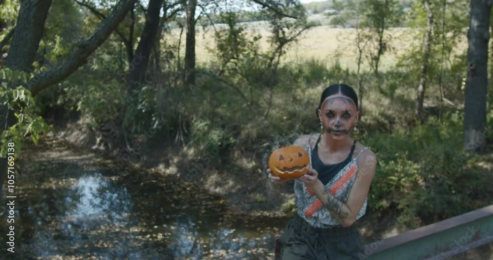Halloween scene as woman in jack-o-lantern facepaint holds up a small jack-o-lantern with white smoke coming out of its face. She is sitting on a bridge railing in a rural area Halloween scene as woman in jack-o-lantern facepaint holds up a small jack-o-lantern with white smoke coming out of its face. She is sitting on a bridge railing in a rural area
