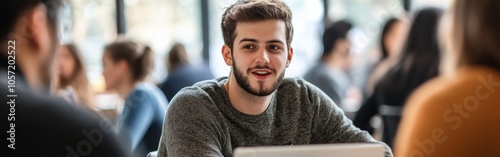 A young man in a sweater talks with friends while using a laptop in a bustling coffee shop filled with patrons at midday