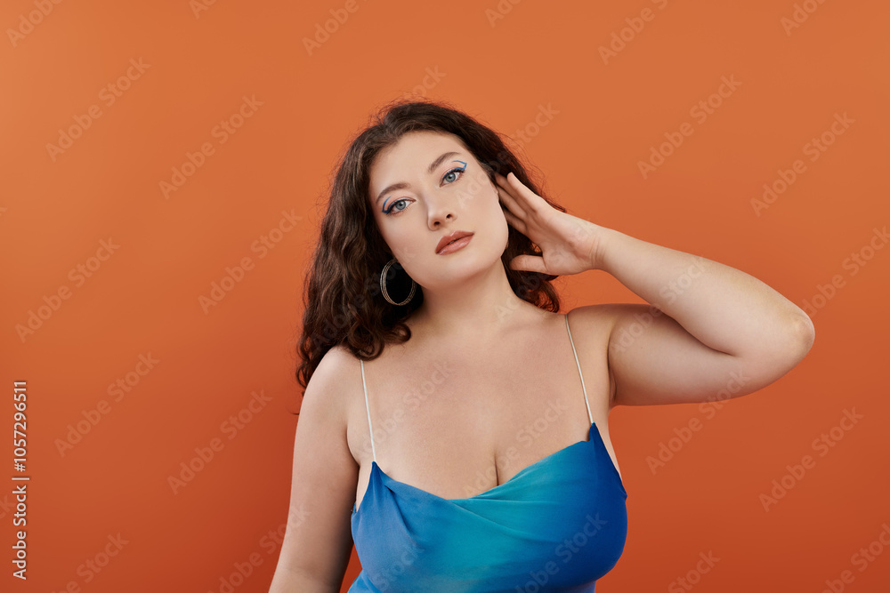 A young, beautiful plus size woman exudes confidence in her blue dress while posing on a bright orange background.