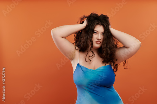 A vibrant young woman in a blue dress with makeup poses confidently on a bold orange background.