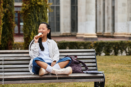 A cheerful young woman eating sandwich while sitting on a bench at a university campus.