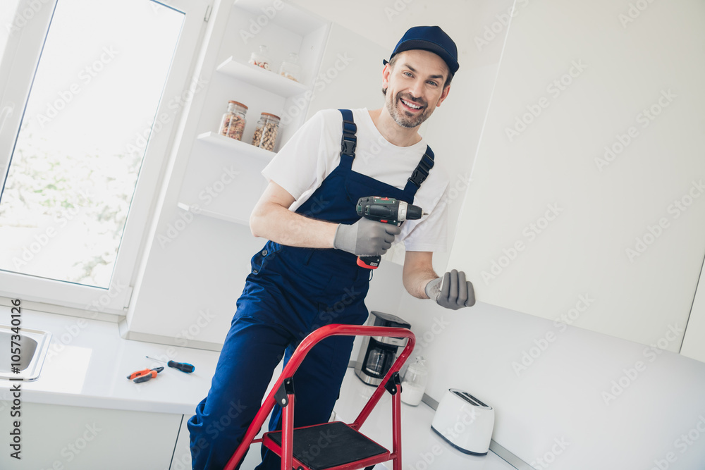 Smiling handyman in overalls using a drill for kitchen renovation and repair work in a modern apartment Smiling handyman in overalls using a drill for kitchen renovation and repair work in a modern apartment