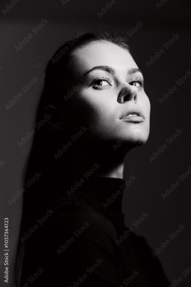 Confident woman with long hair in a black turtleneck, gazing upward against a dark background, embodying strength and elegance. Artistic monochrome portrait.