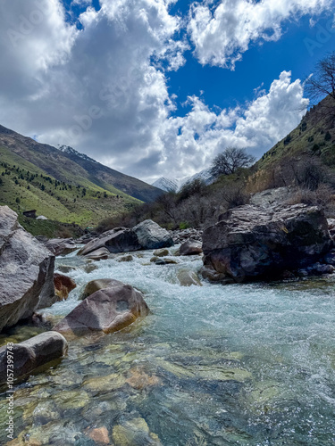 A mountain river in the Alamedin Gorge, Kyrgyzstan, with clear water, surrounded by large boulders and green slopes under a cloudy sky photo