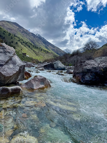 A mountain river in the Alamedin Gorge, Kyrgyzstan, with clear water, surrounded by large boulders and green slopes under a cloudy sky photo