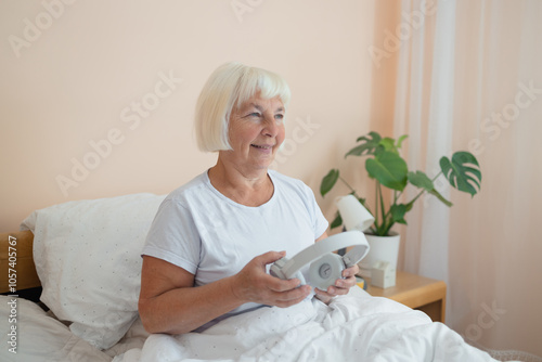 Elderly woman wearing headphones lying on bed using smartphone, embracing technology and enjoying a peaceful moment in her bedroom. 