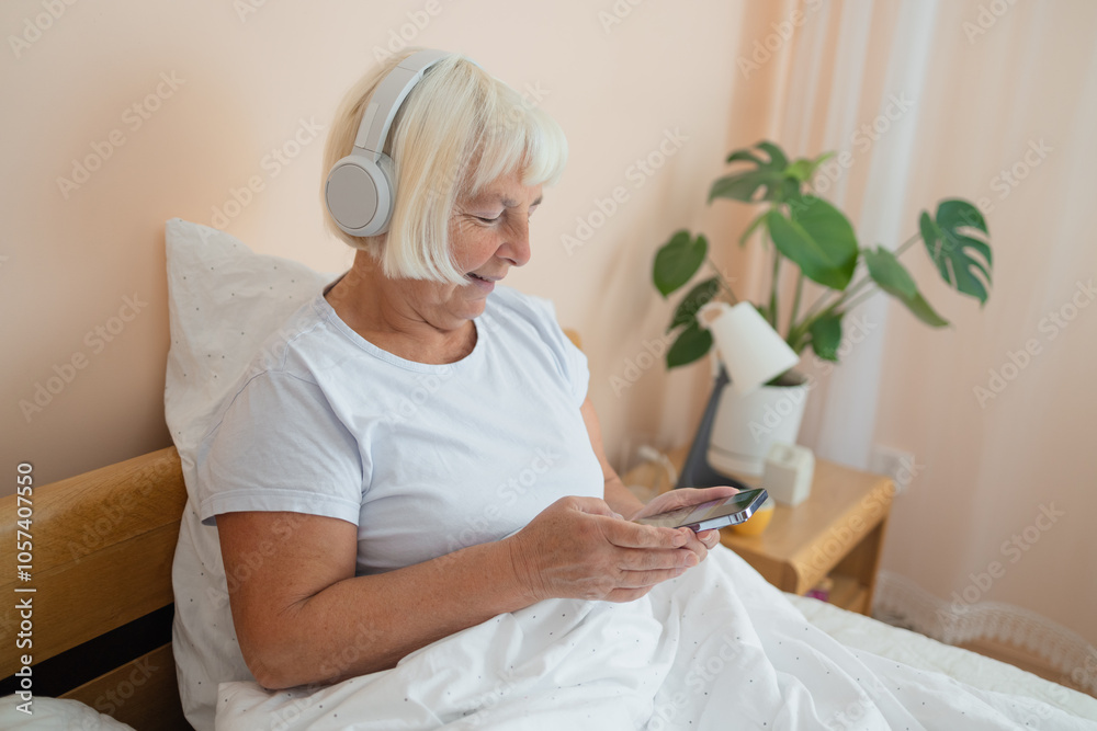 Elderly woman wearing headphones lying on bed using smartphone, embracing technology and enjoying a peaceful moment in her bedroom. Elderly woman wearing headphones lying on bed using smartphone, embracing technology and enjoying a peaceful moment in her bedroom.