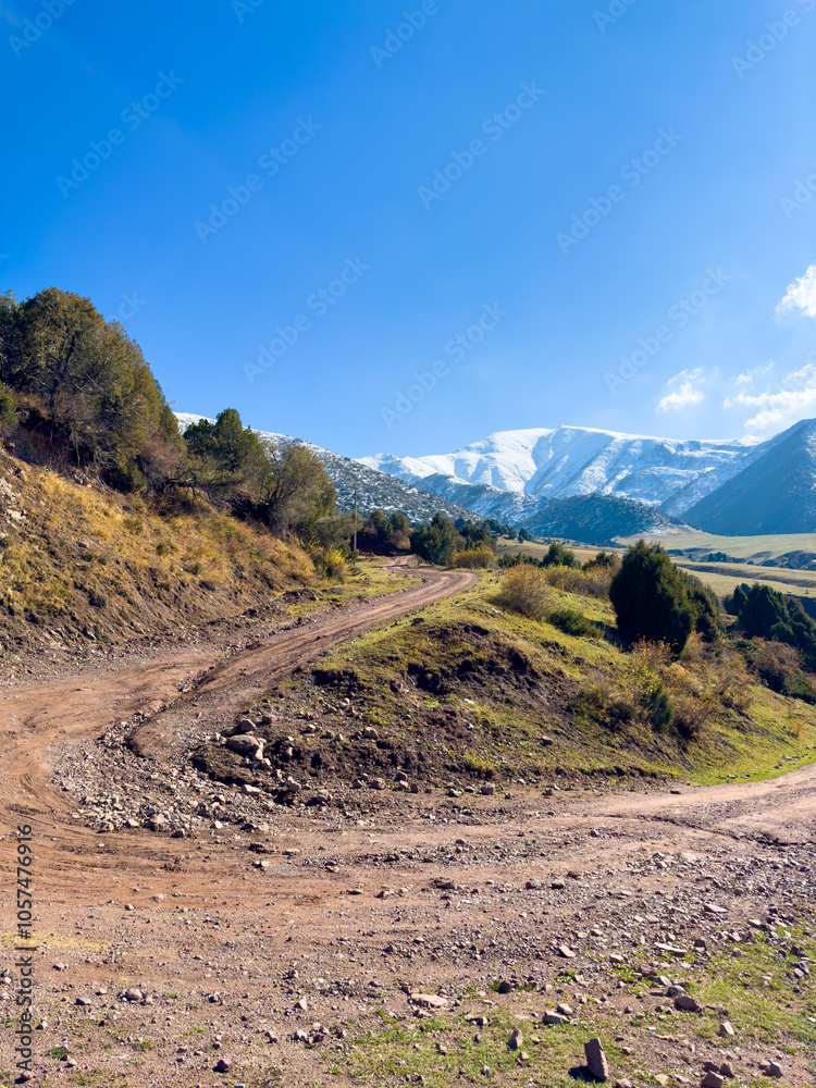 A dirt road winding through the mountain slopes in Kyrgyzstan, with snow-capped peaks in the background and a clear blue sky. A dirt road winding through the mountain slopes in Kyrgyzstan, with snow-capped peaks in the background and a clear blue sky.