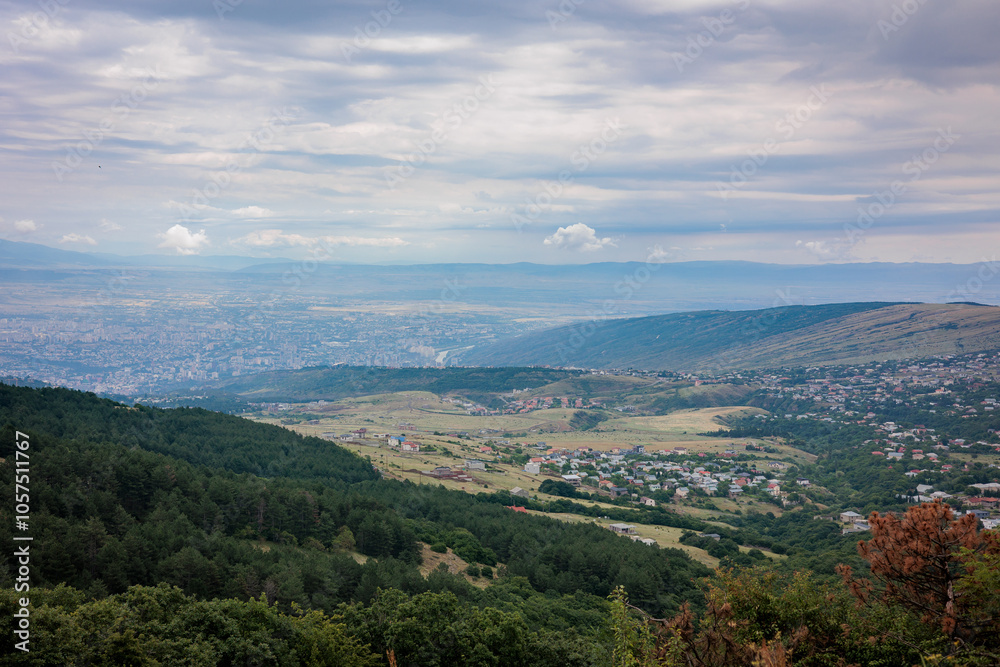 Obraz premium Summer mountain view of Tbilisi, Georgia. Soft clouds cast gentle light over the city and its green landscape, with lush trees in the foreground, creating a calm, scenic atmosphere