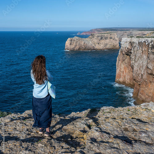 Young woman stands looking out over the beautiful rugged coastline at Sagres Point, location of the Lighthouse of Cabo de Sao Vicente, Algarve, Portugal; Sagres, Faro, Portugal photo