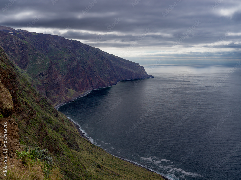 Rugged coastline of the island of Madeira, with a cloudy sky reflecting on the tranquil water of the North Atlantic Ocean, Portugal; Madeira, Portugal