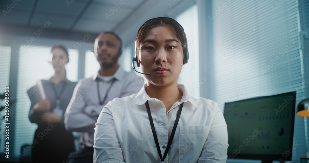 Modern call center control room: Close up portrait of serious Asian female hotline operator in headset looking at camera. Team of technical customer support specialists standing in the background. Modern call center control room: Close up portrait of serious Asian female hotline operator in headset looking at camera. Team of technical customer support specialists standing in the background.