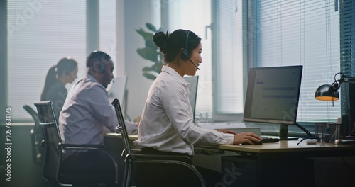 Multiethnic team of technical support operators working in modern call center office. Asian female helpdesk specialist talks on headset, uses computer, looks at camera providing customer service. photo