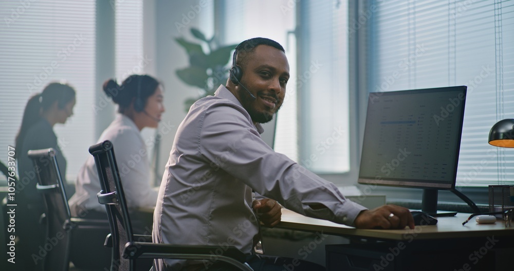 African American technical support operator in headset, sits in front of computer, looks at camera providing customer service. Team of diverse helpdesk specialists work in modern call center office. African American technical support operator in headset, sits in front of computer, looks at camera providing customer service. Team of diverse helpdesk specialists work in modern call center office.