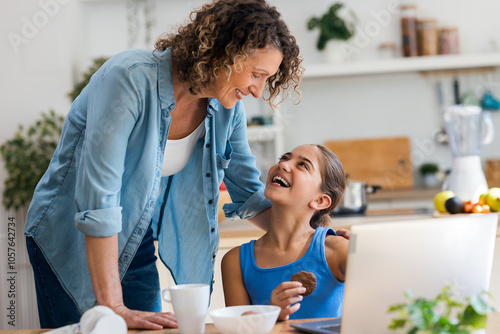 Cute little girl doing homework with laptop with her motherwhile having breakfast in the kitchen photo