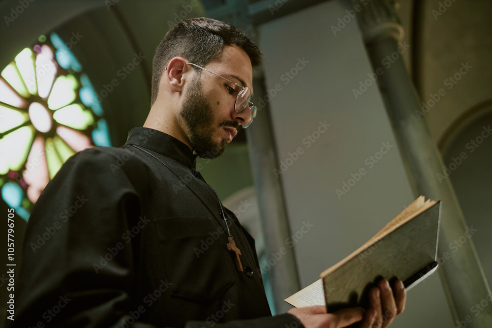 Fototapeta premium Low angle shot of young pastor with glasses and dressed in robe with wooden cross on chest reading Bible during service in local church