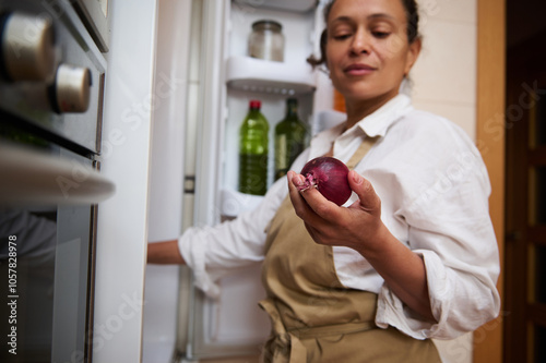 Woman in kitchen holding a red onion while standing by an open refrigerator, wearing a white shirt and apron, captures a moment of cooking preparation and thoughtful meal planning. photo