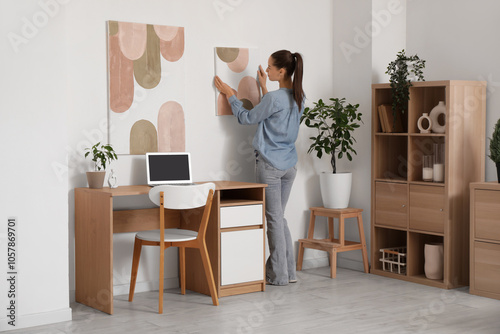 Young beautiful woman hanging picture on white wall near workplace in stylish room