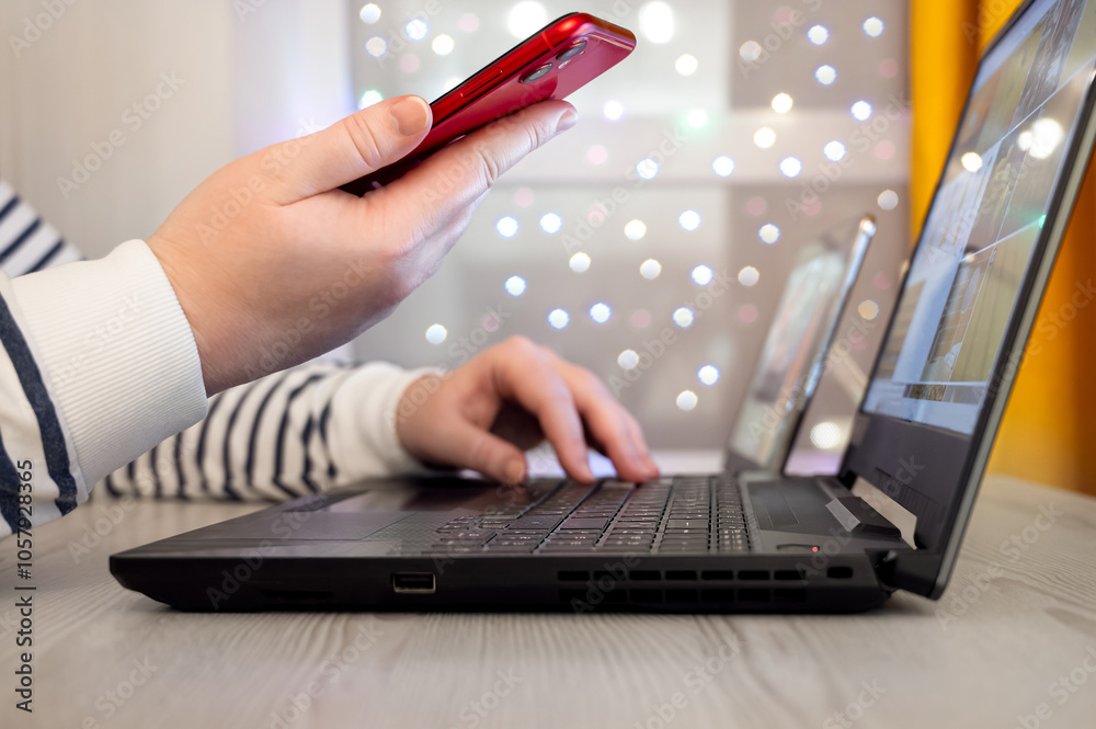 A woman holding a smartphone while working and studying on a laptop creates a cozy atmosphere against a blurred background of festive lights
