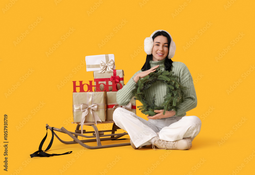Pretty young woman with Christmas wreath, sledge and gift boxes sitting on yellow background Pretty young woman with Christmas wreath, sledge and gift boxes sitting on yellow background