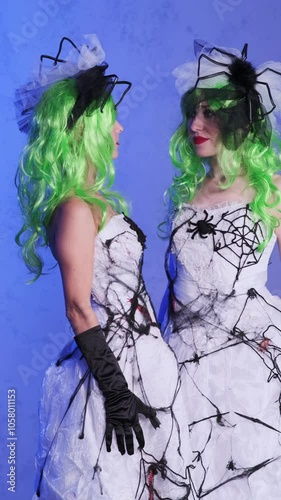 Two girls in scary dresses with evening web wedding, green hair and head decorations look at each other on a blue background. Scary bride costume in sight. The celebration was open. Dead bride costume