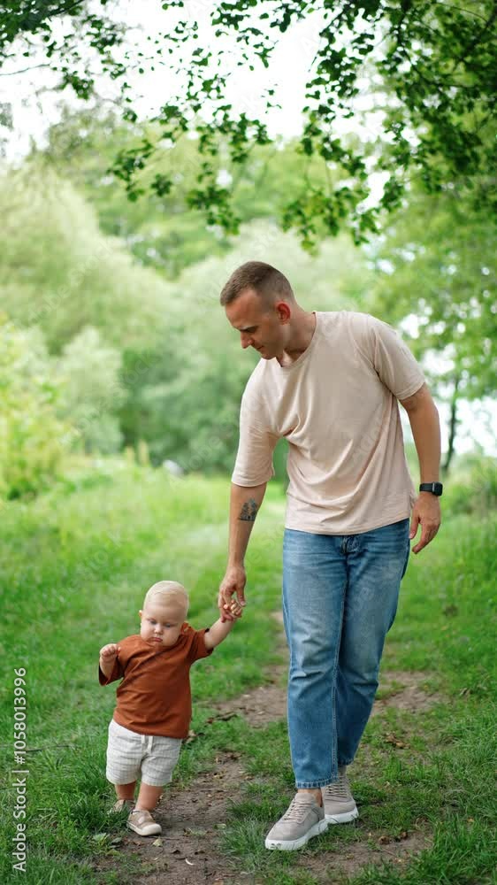 Father holds his little son by the hand walking by the park. Teaching a child to walk outdoors. Vertical video. Father holds his little son by the hand walking by the park. Teaching a child to walk outdoors. Vertical video.