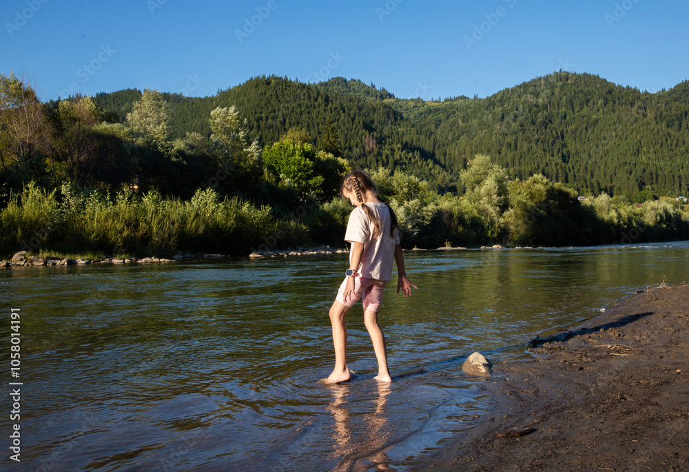 Fototapeta premium A girl stands in shallow water, feeling the cool river flow around her feet while sunlight filters through surrounding trees, creating a serene atmosphere filled with nature's beauty.