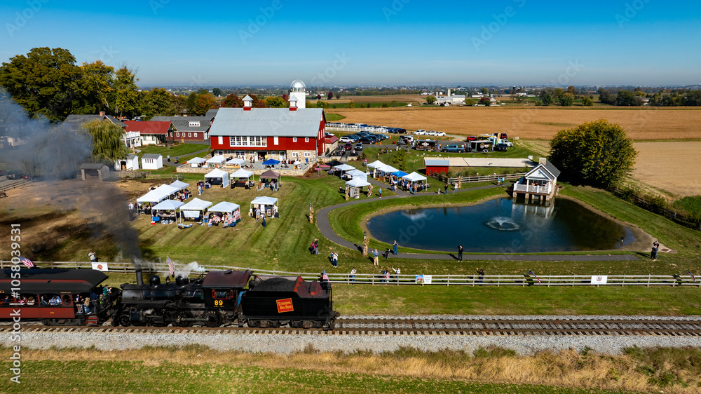 Fototapeta premium A steam train travels along the tracks near a bustling rural fair, with families gathered around tents, enjoying activities. A serene pond and farm buildings are visible under a bright blue sky.