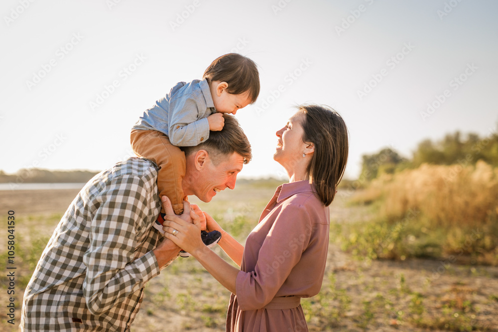 Fototapeta premium Happy family walking on sandy beach of river. Father, mother holding baby son on hands and going together. Rear view. Family Ties concept.