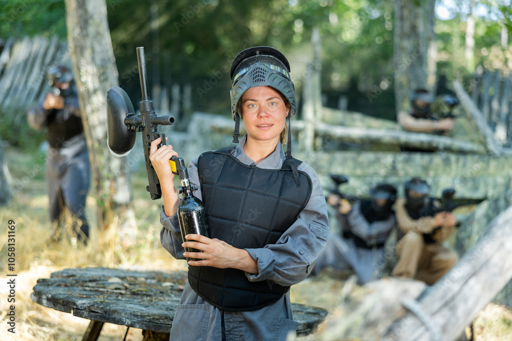 Fototapeta premium Smiling woman wearing uniform and holding gun ready for playing paintball with friends outdoor