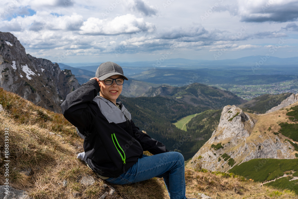 Smiling boy sitting on mountainside with scenic view of the valley in the background on cloudy day. Concept of adventure, nature exploration and outdoor recreation at high altitude. Smiling boy sitting on mountainside with scenic view of the valley in the background on cloudy day. Concept of adventure, nature exploration and outdoor recreation at high altitude.