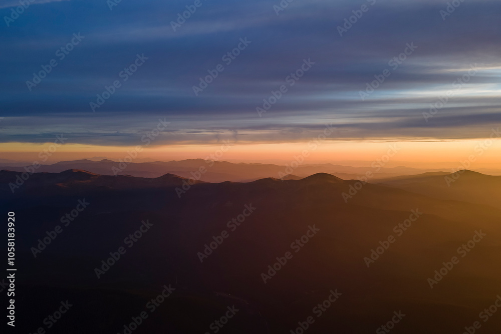 Aerial view of dark mountain hills with bright sunrays of setting sun at sunset. Hazy peaks and misty valleys in evening